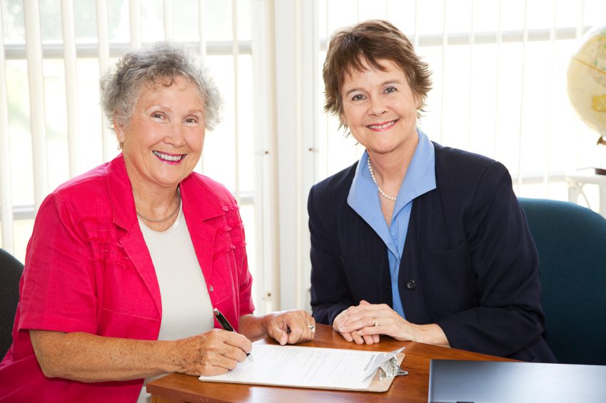 Senior woman signs paperwork in mature businesswoman's office.